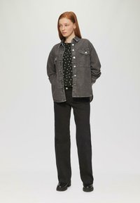 Zwart denim shirt met knoopsluitingen en zakken, gelaagd over een zwart top met stippen. Gecombineerd met zwarte rechte broek en zwarte schoenen.