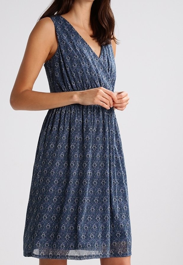Robe bleue sans manches longueur genou avec un motif géométrique subtil, taille cintrée et décolleté en V portée par une personne debout les mains jointes.