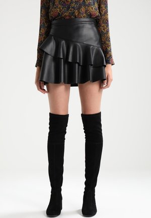 Kobieta w czarnej skórzanej, falbaniastej mini spódnicy, kwiecistej bluzce z długim rękawem oraz czarnych zamszowych kozakach za kolano, stojąca na jednolitym tle.