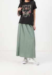 T-shirt grafico nero con design floreale, abbinato a una lunga gonna verde fluente e sneakers bianche alte. Borsa in pelle semplice a spalla.