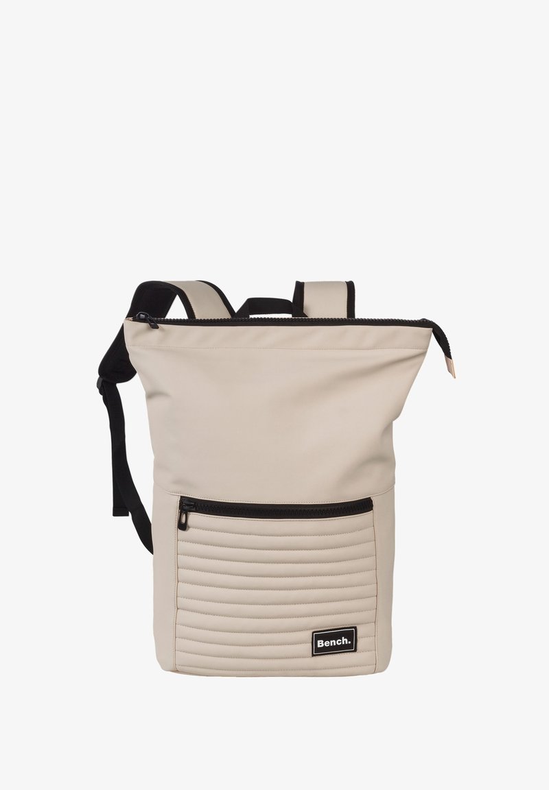 Beige rugzak met een ritssluiting aan de bovenkant, een getextureerd paneel aan de onderkant met verticale ribbels, verstelbare zwarte riemen en een voorvak met rits.