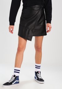 Svart läderwrapkjol med sned skärning och spänndetalj, i kombination med marinblå och vita hög-top sneakers och randiga strumpor.