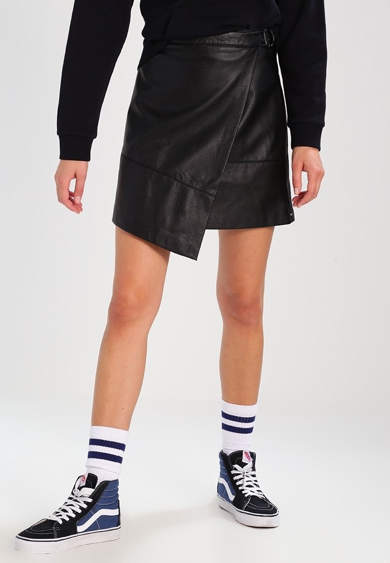 Svart läderwrapkjol med sned skärning och spänndetalj, i kombination med marinblå och vita hög-top sneakers och randiga strumpor.