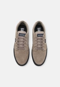 Tan mockasiner i suède med rund tå, mörk gummisula och beiga snören. Har en svart innersula med vit logotyp på sidan och tungan.