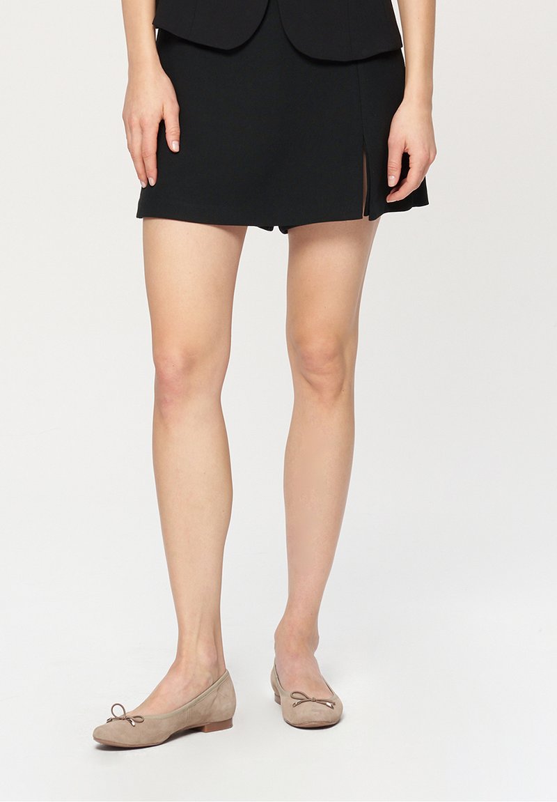 Czarna mini spódnica z bocznym rozcięciem, zestawiona z beżowymi zamszowymi balerinkami z małym akcentem kokardki na palcu. Gładka tekstura całości.
