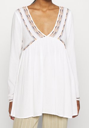 Blouse blanche à manches longues avec un décolleté en V profond et une bordure décorative cousue, portée avec un pantalon beige à rayures.