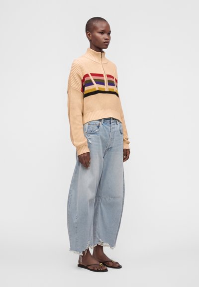 Personne portant un pull en maille beige avec des rayures colorées, un jean large bleu clair délavé et des sandales noires, debout sur un fond uni.