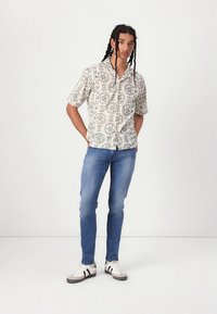 Košulja kratkih rukava svijetlo bež boje s tamnim uzorkovanim naglascima, uparena s svijetloplavim trapericama i crno-bijelim tenisicama.