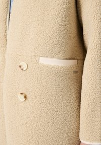 Beige fleece jas met een gestructureerde buitenkant, twee grote knopen en een verborgen zak met een crèmekleurige accentaan de voorkant.