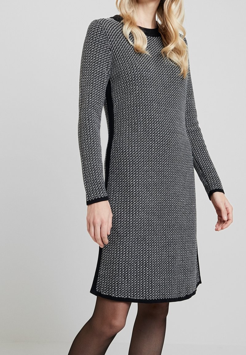 Femme portant une robe longue texturée noir et blanc à manches longues, longueur genou, avec des panneaux latéraux noirs, associée à des collants noirs transparents.