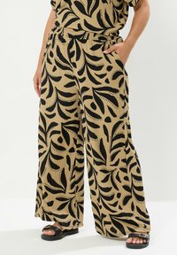 Breda byxor i en beige tyg med ett djärvt svart lövmönster, elastisk midja och fickor, i kombination med svarta sandaler.