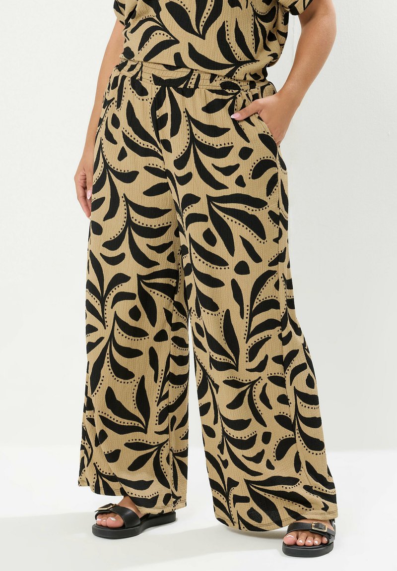 Breda byxor i en beige tyg med ett djärvt svart lövmönster, elastisk midja och fickor, i kombination med svarta sandaler.