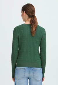 Pull en laine vert à mailles torsadées, à manches longues, avec un col rond et un ourlet côtelé, associé à un jean bleu clair. La texture montre des motifs complexes.