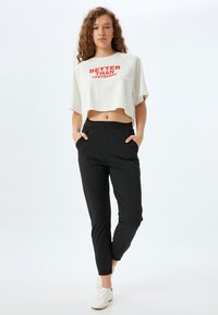 Camiseta blanca recortada con texto rojo, combinada con pantalones jogger de cintura elástica negros. El modelo lleva zapatillas blancas y tiene las manos en los bolsillos.