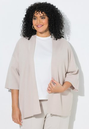 Kvinde med krøllet sort hår, smilende, iført guld øreringe i hoop-stil, en hvid top og en lys beige cardigan med åben front samt bukser.