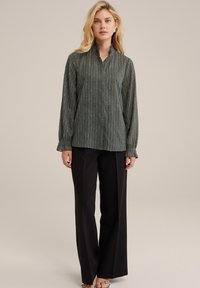 Zöld, hosszú ujjú blúz függőleges csíkokkal és mandarin gallérral, fekete, széles lábú nadrággal és átlátszó sarkú szandállal párosítva.