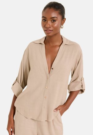 Frau trägt beiges, geknöpftes Hemd mit hochgekrempelten Ärmeln und passende Hose, posiert mit einer Hand in der Tasche vor weißem Hintergrund.