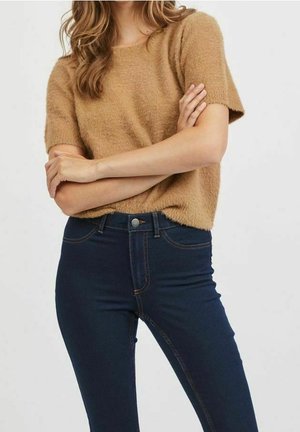 Vrouw draagt een korte mouw fuzzy trui in kamelkleur en donkerblauwe high-waisted skinny jeans met gekruiste handen over de borst.