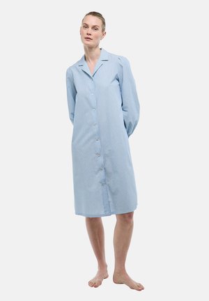 Femme debout pieds nus portant une robe chemise bleu clair à manches longues, boutonnée jusqu'au genou, avec de subtiles rayures verticales.
