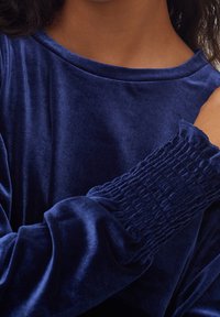 Marinblå sammetstopp med en mjuk textur, med en båturringning och smockdetaljer vid ärmarna för en samlad effekt.