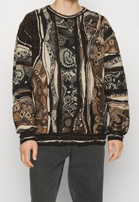 Sweter dziany z wzorem patchworkowym w ziemistych odcieniach brązu, czerni i kremu oraz złożonymi geometrycznymi wzorami. Ściągacze na mankietach i dole swetra.