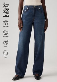 Person med mörkblå vida denimjeans och svarta slip-on-skor, som står med armarna avslappnade längs sidorna mot en ljus bakgrund.