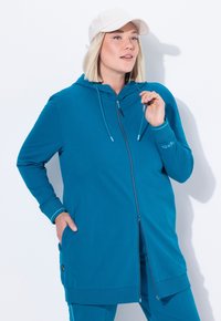 Pitkä turkoosi vetoketjullinen huppari, jossa on huppu, sivutaskut ja ribbireunukset. Korostaa erottuvia ompeluée yksityiskohtia ja pientä logoa hihassa.