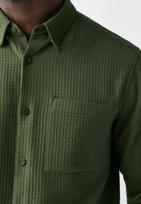 Groene, gestructureerde button-up shirt met een kraag, voorzien van een borstzak en vijf knopen. Het materiaal heeft een wafelachtig patroon.