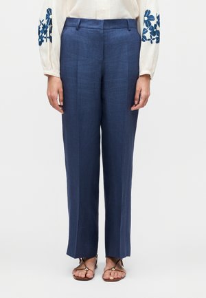 Kvinde iført blå bukser med lige ben, en hvid bluse med blå blomsterbroderi på ærmerne og brune åbne sandaler.