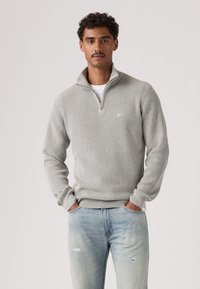 Pilkas ribotuotas megztinis su ketvirčio užtrauktuko iškirpte, ilgomis rankovėmis ir tekstūruota apdaila, derintas su šviesiai mėlynais, nuspaudytais džinsais.