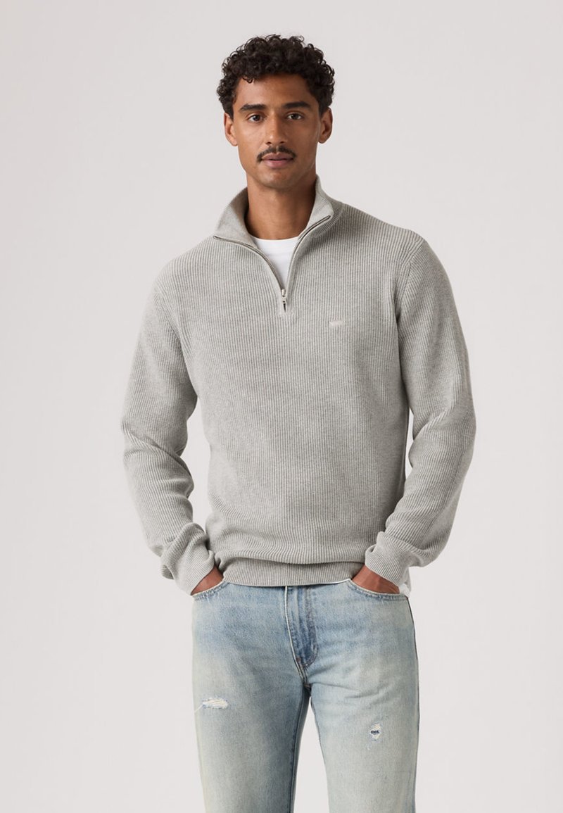 Pull gris côtelé avec un col à fermeture éclair d'un quart, doté de manches longues et d'une finition texturée, associé à un jean bleu clair déchiré.