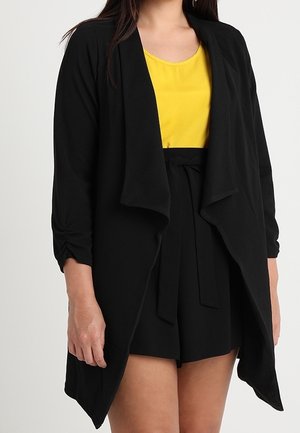 Femme portant un cardigan noir ample ouvert devant, un haut sans manches jaune et un short noir taille haute avec une ceinture nouée à la taille.