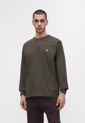 Jonge man met kort haar die een olijfgroene longsleeve draagt met een klein geel logo op de borst en een donkere broek.