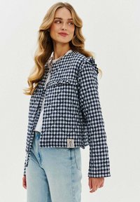 Kurtka w pikowany wzór w granatowo-białą kratę, z falbaniastymi ramionami, dwoma przednimi kieszeniami i naszywką z logo w pobliżu dolnej krawędzi. Lekki materiał.