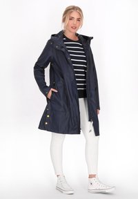Marinblå vattentät jacka med huva, dragkedja och tryckknappar; har två framfickor och en lätt design. Modellen bär vita byxor och sneakers.