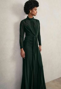Tumed roheline pikkade varrukatega kleit kõrge kaelusega, drapeeritud disainiga ja ette seotud. Sujuv tekstuur ja põrandani ulatuv, kergelt külgsuine lõhiku igas.
