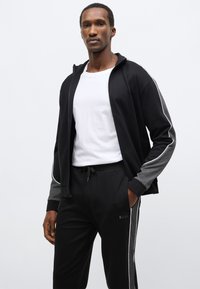 Čierna bunda na zips so sivými doplnkami a bielymi pruhmi na bokoch, doplnená čiernymi nohavicami s vyšitým logom. Zmes bavlny, voľný strih.