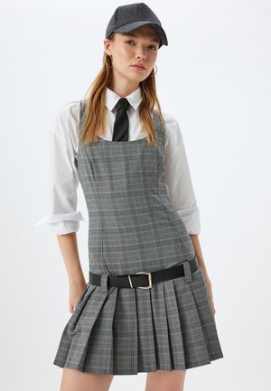 Abito grigio a quadri con un corsetto aderente e una gonna plissettata. Abbinato a una camicia bianca con bottoni e una cravatta nera. Una cintura nera evidenzia il punto vita.