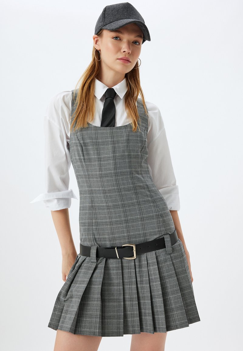 Vestido de cuadro gris con un corpiño ajustado y falda plisada. Combinado con una camisa blanca de botones y una corbata negra. Un cinturón negro acentúa la cintura.