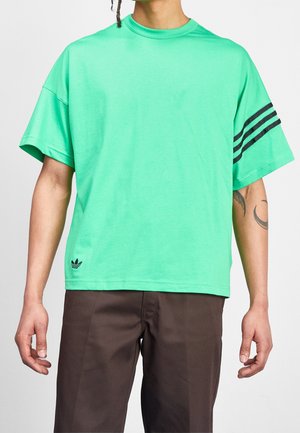 Osoba v volném mátovém tričku Adidas s černými pruhy na jednom rukávu a hnědých kalhotách, viditelné tetování na levém předloktí.