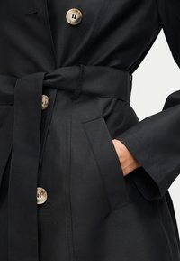 Μαύρο παλτό με μεγάλα μπεζ κουμπιά και φαρδιά ζώνη δεμένη στη μέση, χέρι τοποθετημένο μέσα σε εμπρόσθια τσέπη, κοντινή λήψη.