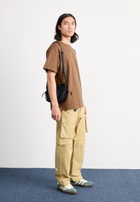 Brun bomull t-shirt, beige cargobyxor med flera fickor och gröna och vita sneakers. Svart påse med dragsko och mesh detaljer.