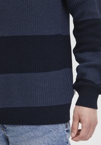 Pulover tricotat cu dungi orizontale bleu marin și albastru închis, având manșete și tiv ribbed, amplasat lângă blugi din denim deschis.