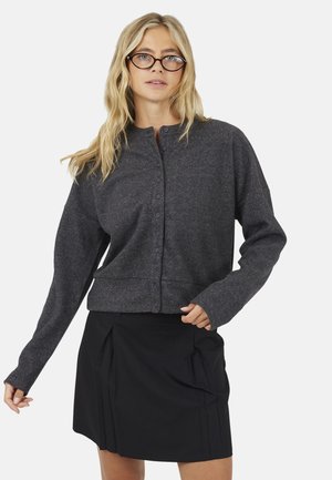 Blondine vrouw draagt een donkergrijs vest met knopen en een zwarte rok, met een bril, staand tegen een effen lichte achtergrond.