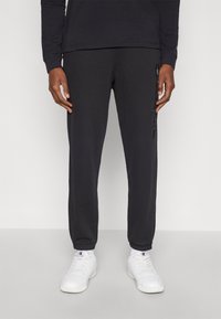 Μαύροι φόρμες με κορδόνι, στενό τελείωμα στα πόδια, ριμπ μανσέτες και διακριτικό λογότυπο στο πλάι. Συνδυάζονται με λευκά αθλητικά παπούτσια και ορατές κάλτσες αστραγάλου.