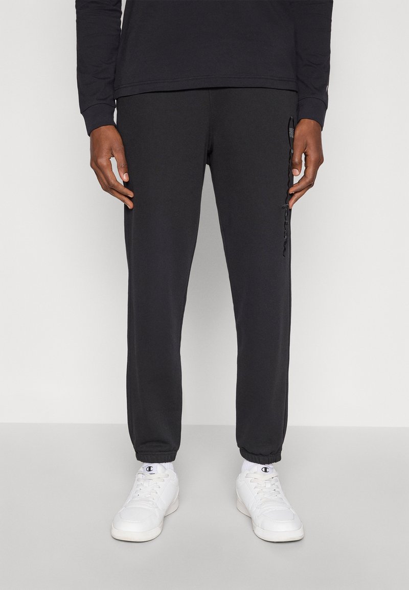 Μαύροι φόρμες με κορδόνι, στενό τελείωμα στα πόδια, ριμπ μανσέτες και διακριτικό λογότυπο στο πλάι. Συνδυάζονται με λευκά αθλητικά παπούτσια και ορατές κάλτσες αστραγάλου.