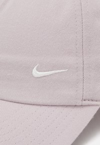 Gaiši purpura beisbola cepure, kas izgatavota no teksturēta auduma, ar izliekto malu un mazu balta izšūtu Nike logo priekšpusē.