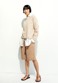 Gestreifter beigefarbener und weißer übergroßer Pullover, der über einem weißen Hemd getragen wird, kombiniert mit beigen Shorts und schwarzen Sandalen mit roten Akzenten.