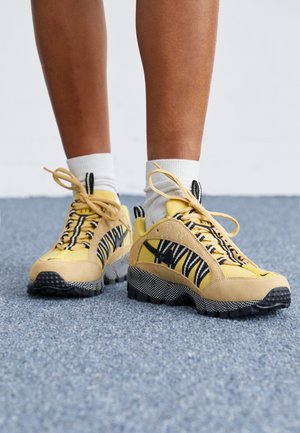 Gule ruskinds- og mesh-sneakers med sorte og hvide stribede detaljer, strukturerede såler og snørebåndsdesign. Hvide sokker synlige over anklen.