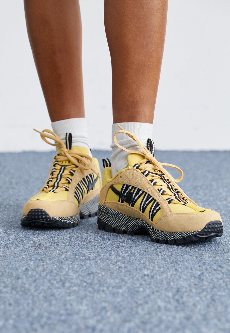 Des baskets en suédine jaune et en mesh avec des accents rayés noirs et blancs, des semelles texturées et un design à lacets. Des chaussettes blanches visibles au-dessus de la cheville.